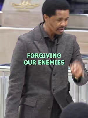 A man in a blazer is captured from the torso up, centered within the frame. The background appears to be a neutral-toned interior, possibly a stage or auditorium. Overlayed on the image is a text block in a light green color, reading 'FORGIVING OUR ENEMIES'. The man is looking slightly to the right, possibly addressing an audience. The lighting seems to be even, without strong shadows, and the overall tone appears to be serious and reflective.
