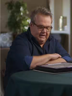 The image shows a man wearing glasses, seated at a table covered with a dark green tablecloth. He's looking downward with a focused expression. The background is slightly blurred. The lighting is warm and natural.