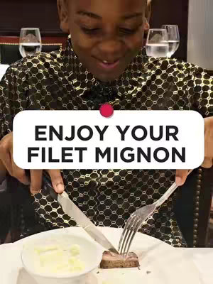 A person is shown smiling and cutting a piece of steak at a fancy restaurant. The individual is wearing a dark patterned shirt sitting at a table with silverware and plates. The overlay text reads 'Enjoy Your Filet Mignon'.
