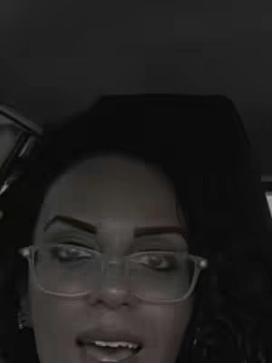 A close-up, monochromatic shot of a woman's face. She is wearing glasses and appears to be in a vehicle; only the upper part of her body is visible. The background is dark.