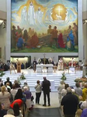 The image captures a wide shot inside a large, well-lit church or cathedral. The focus is on a religious ceremony taking place at what appears to be a high altar. In the background, a large mural depicting biblical scenes dominates the upper portion of the image. Beneath the mural, the altar is set with religious objects, and a priest in white and gold vestments is seen officiating. A crowd of people, likely attendees, are visible in the foreground, facing the altar. The overall composition gives a sense of solemnity and reverence associated with religious practices.
