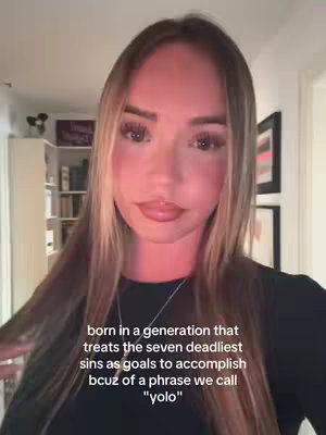 A woman with long, straight blonde hair is centered in a selfie-style shot, looking directly at the viewer. She has full lips, and eye makeup is visible. Behind her, a blurred background suggests a domestic setting, with shelves and wall decorations. The lighting is soft, and it appears indoors, with warmer color casts.