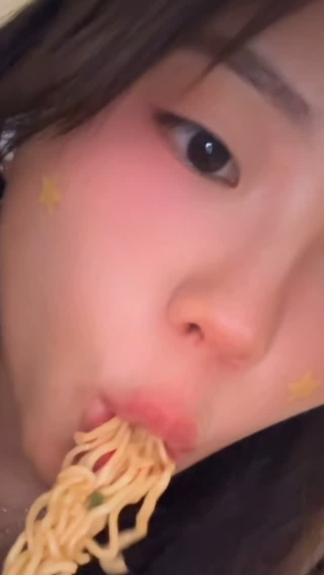 A close-up shot features a person eating noodles. Their visible portion emphasizes an eye with a small star sticker, a cheek with blush, and lips. Noodles are pulled into the mouth.