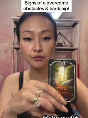 A woman with dark hair and makeup holds a tarot card towards the camera. She's in front of a blurred pink background. The tarot card shows a path through trees. Text at the top says 'Signs of a overcome obstacles & hardship!'.