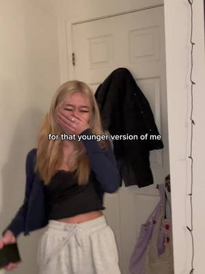 A young woman with long blonde hair is covering her mouth with her right hand, seemingly laughing or hiding a smile. She is wearing a black top paired with a navy blue cardigan and light sweatpants. The background includes a white door with a black jacket hanging on it.