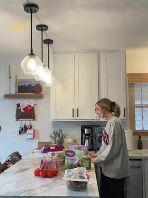 A woman is in a kitchen, standing at a marble countertop. There is food laid out. She stands in front of a counter space with items such as the kitchen sink, a coffee machine, and products. The kitchen has white cabinets, pendant lights, and a window.