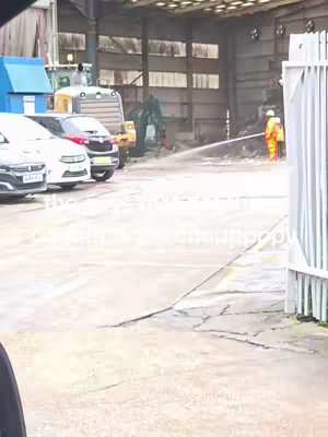 An outdoor scene presents a parking lot with several cars parked parallel on the asphalt. Beyond the vehicles, large buildings with metal siding and various machinery are visible. In the background, a person in an orange safety suit is using a hose to spray water, with a visible stream extending towards the left. The overall lighting is overcast and somewhat dim, casting shadows and a hazy atmosphere.