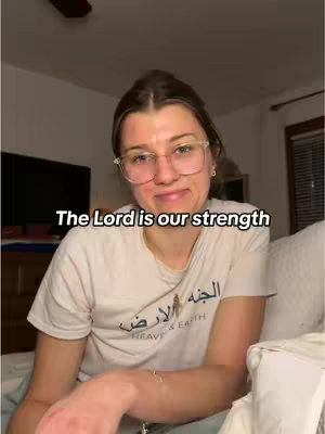 A woman with glasses and a t-shirt is sitting on a bed, looking at the camera. Text overlay in black on the top-left corner with bold font that reads "The Lord is our strength."