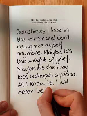 An open book on a wooden surface. The left page contains handwritten text in black ink with the beginning of a sentence in the corner. The hand of the writer is seen holding a pen at the bottom of the page.