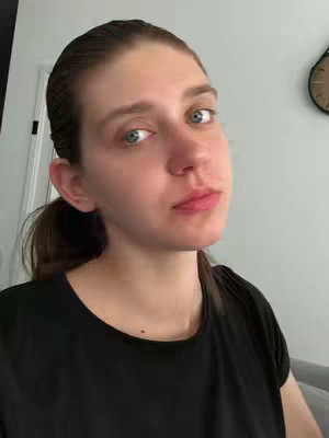 A woman with light skin and brown hair pulled back into a low ponytail looks directly at the camera, with a slight smirk. She is wearing a black shirt. The background is a room with a white wall and a visible clock.