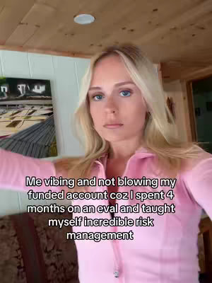 A selfie-style video with a young woman as the focal point. She has blonde hair, fair skin, and is wearing a pink collared shirt. The text overlay indicates it's about trading and achieving funded accounts. The background appears to be a room with wood paneling and a painting hanging on the wall.