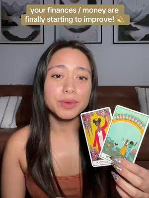 A woman with long dark hair and in a casual outfit is holding up two tarot cards in her hands. The background shows a blurred, warm-toned interior with three framed pictures on the wall. Above her, a text overlay reads, "your finances / money are finally starting to improve!"