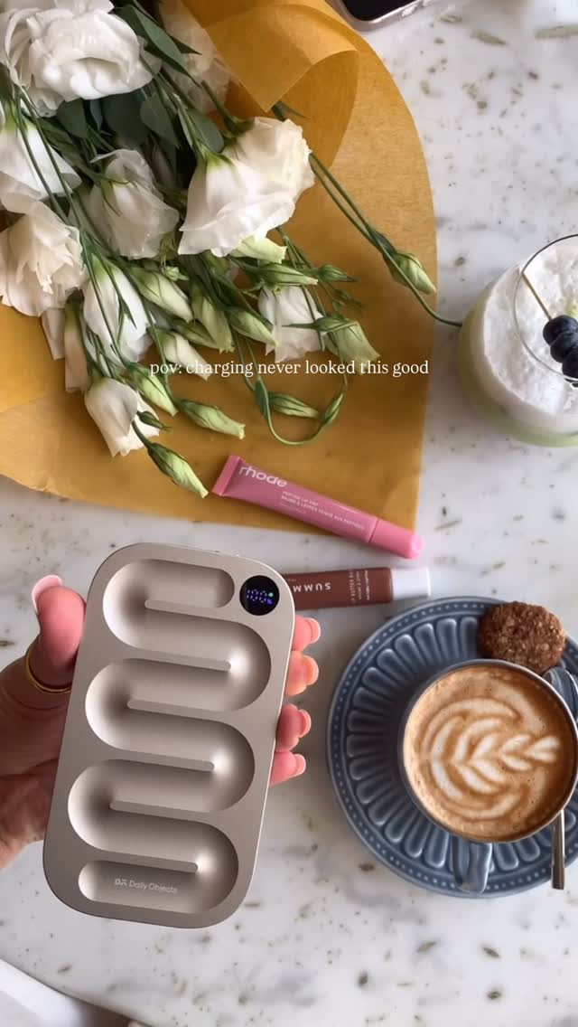 A top-down shot captures a still life arrangement with a bouquet of white flowers in a yellow paper wrap on the top left. Below that, a gold and white charging device is being held by a hand with pink nail polish. Various beauty products and a cup of coffee with latte art are also present, all set against a marble surface.