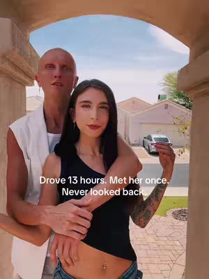 A couple stands embracing each other within an arched doorway. The man, with a shaved head and white sleeveless shirt, wraps his arms around the woman. The woman has long, dark hair, and a black top. The background shows a residential street and blue sky.