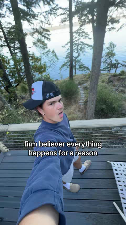 A young man in a backward baseball cap stands on a wooden deck with a scenic view of a lake and trees. He is wearing a blue sweatshirt, white shorts, and beige shoes, looking directly at the camera with an open mouth expression. Text overlay at the bottom reads a motivational quote.