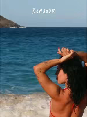 A woman with dark curly hair stands on a beach, facing away from the camera, with the ocean in the background. She is wearing a red bikini top and has her arms raised, with her hands touching her head. The word "BONJOUR" is written at the top of the image.