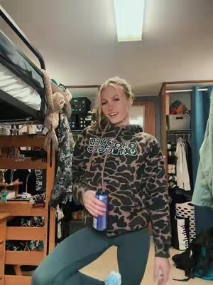 A young woman with blonde hair is in a dorm room. She is wearing a patterned hoodie, leggings, and holding a can. The room is cluttered, with a bunk bed in the background.