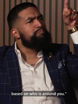 A man is the focus, with a composed expression and a finger pointing up. He has a well-groomed beard, a white shirt, and a blue plaid blazer. A gold watch and ring are visible. The background is a neutral color with vertical lines.