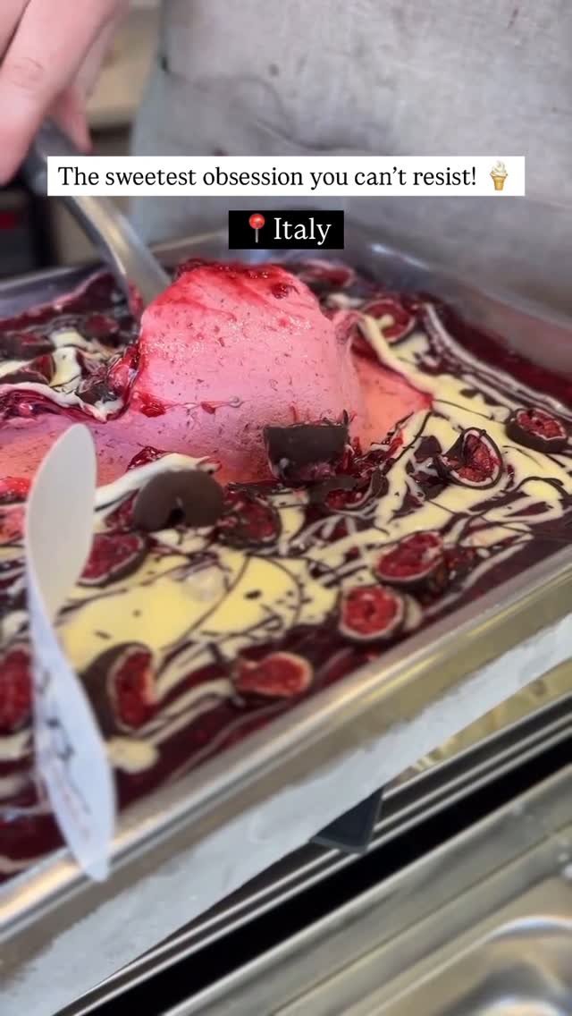 A close-up shot of a silver metal tray filled with gelato. The gelato has a pink base with lighter swirls and ribbons of dark red or maroon running through it; some have a white creamy element. There are also pieces of chocolate and what appears to be fresh berries half-dipped in chocolate scattered throughout the gelato. On the left, a white spoon is partially visible dipping into the gelato.