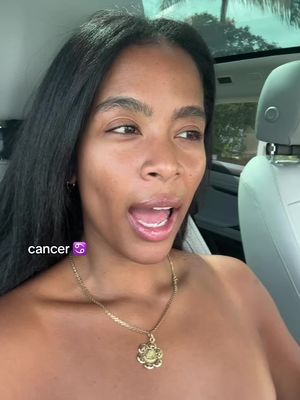 A woman with dark hair and a gold necklace is seated inside of a car. She is looking to the side with an open mouth and talking. A graphic of "cancer" is shown in the bottom left corner.