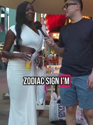 A woman in a white dress is being interviewed on the street by a man wearing glasses, both holding microphones. The background shows what appears to be a shop or storefront and red decorations, with the text overlayed on the bottom half of the image.