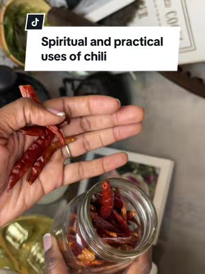 A person's hands hold a jar of dried chili peppers and a handful of the peppers. Overlay text in a white box reads "Spiritual and practical uses of chili". The background is blurred with other kitchen items.