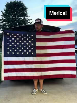 A man stands holding a large American flag with the stars and stripes, obscuring his body from the waist up. He is wearing a baseball cap and sunglasses. In the top right corner, a stylized blue box contains the word "Merica!". The background features a tree and a sky.