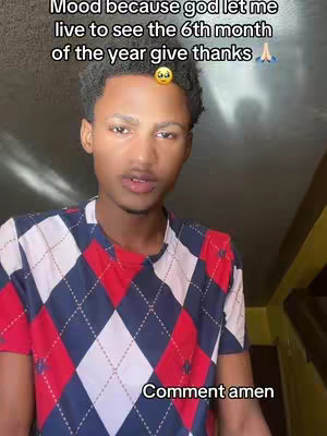 A young man with short, dark hair is seen from the chest up, in a selfie-style shot. He's wearing a shirt with a red, white, and blue argyle pattern. Above his head is overlaid text in a sans-serif font on a dark background; the text says, "Mood because god let me live to see the 6th month of the year give thanks". Above the text and slightly to the right, there is a small image of a sad face emoji. In the bottom of the frame is the words, 'Comment amen' also in sans-serif font.