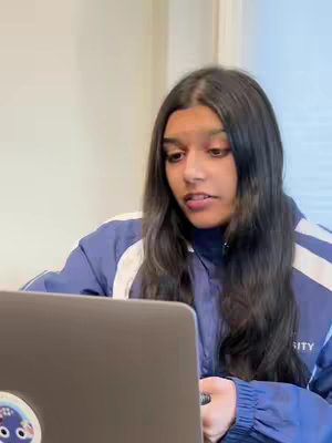 A woman with long black hair in a blue jacket is looking down at a laptop with a sticker on it.