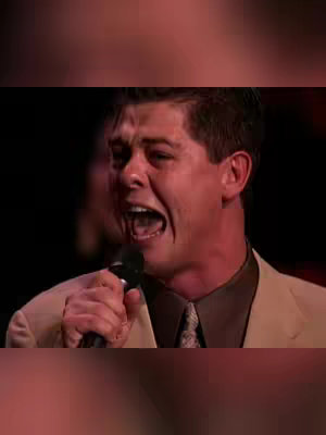 A close-up shot of a man with short, dark hair and a light-colored suit, holding a microphone and singing with a passionate and emotive expression; his mouth is wide open as if belting out a note, and his eyes show intensity. The lighting is focused, and the background is blurred, suggesting a performance or concert setting.