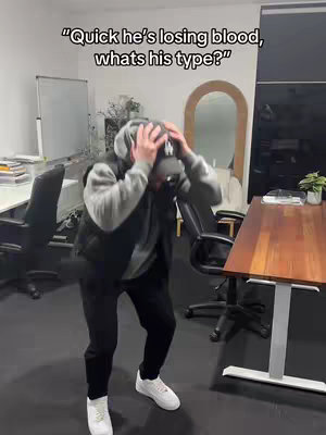 A man is in an office setting, covering his head with his hands, appearing distressed. Text overlays read "Quick he's losing blood, what's his type?" hinting at a dramatic or comedic scenario.