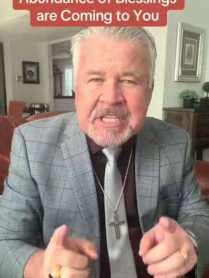 A middle-aged man with gray hair and a beard, wearing a gray plaid suit with a burgundy shirt and a silver tie. He is wearing a cross necklace and is pointing towards the viewer with both hands. The background features a living room setting.