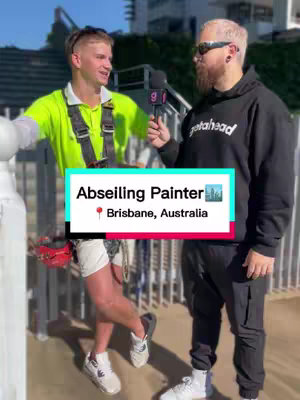 Two men are standing outside, near a railing with a brightly colored backdrop. The man on the left is wearing a neon green shirt, work gear, and a harness, while the man on the right is wearing a black hoodie and sunglasses and is holding a microphone. A text overlay provides the video's title.