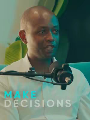 A close-up shot of a man, likely in a studio setting, wearing a white shirt and speaking into a microphone. The background has a teal color. Overlayed text at the bottom says 'MAKE DECISIONS'.