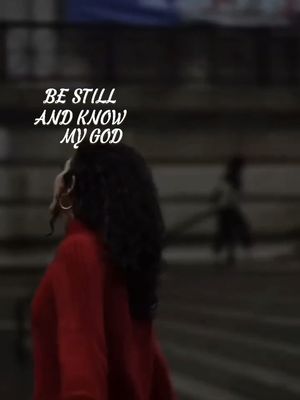 A woman in a red sweater is captured from the back, looking up with a thoughtful expression. Overlaid on the image is white text reading "BE STILL AND KNOW MY GOD." The background is blurred, suggesting an indoor or urban setting.