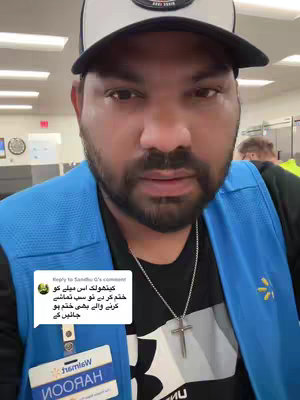 A man with a beard and a black cap is looking towards the camera. He is wearing a blue vest with a Wal-Mart logo on it. A cross necklace is seen dangling around his neck, and the background is a Wal-Mart setting.