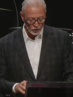 A medium shot of a well-dressed, older man wearing glasses and a white shirt with a blazer. He is looking down and speaking, with an apparent microphone in front of him. The background is dark.