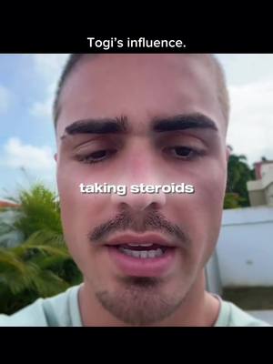 A selfie-style shot featuring a young man in a close-up view. The man has short hair, dark eyebrows, and a mustache. The background is a blurred outdoor setting with green foliage and a blue sky. Text overlayed on the image reads 'taking steroids'.
