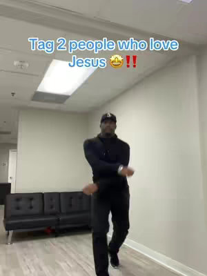 A man wearing a black outfit and a baseball cap is dancing in an office-like space. Overhead, a fluorescent light illuminates the scene. The image features text overlayed at the top.