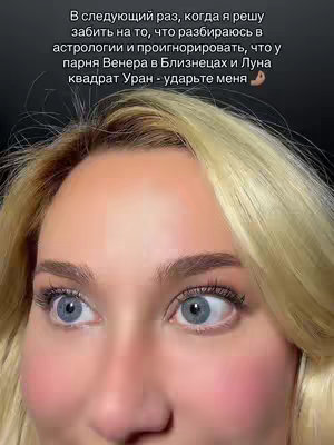 A close-up shot of a light-skinned woman with blonde hair partially covering her forehead, staring wide-eyed towards the left of the frame. Her eyes are very blue with prominent lashes, and her cheeks are flushed a light pink. The background is dark, creating a focus on her face. Text in Russian is overlaid above her head.