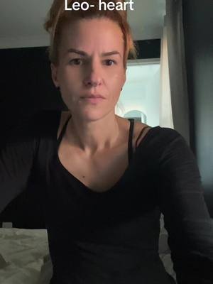 A woman in a black top looks directly at the camera. The words "Leo-heart" are above her head. The background is dark with glimpses of a window and curtain.