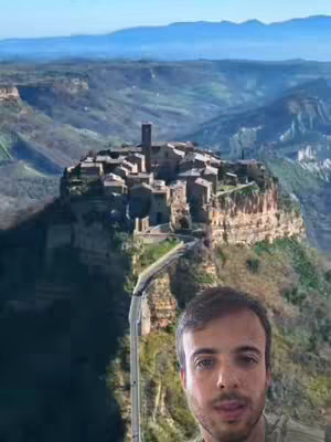 The cover image shows a man's face overlaid on an aerial shot of a historic town nestled on a cliff. The town is made up of stone buildings, and a road leads up to the cliff. Rolling hills and distant mountains fill the background, under a blue sky.