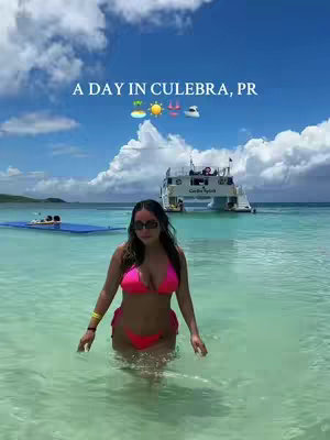 A woman in a pink bikini stands in clear turquoise water, facing the camera. Behind her are a boat and a beautiful blue sky with fluffy white clouds. Text overlay reads, 'A Day in Culebra, PR'.