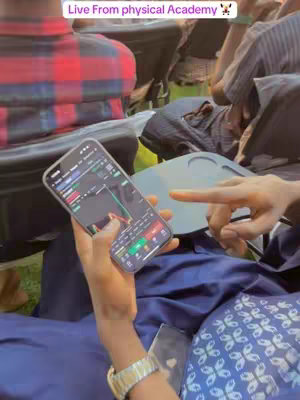 A person holds a smartphone displaying financial data overlaid with the text "Live From physical Academy". The person wears a wristwatch and sits in a casual outdoor setting, surrounded by other people.