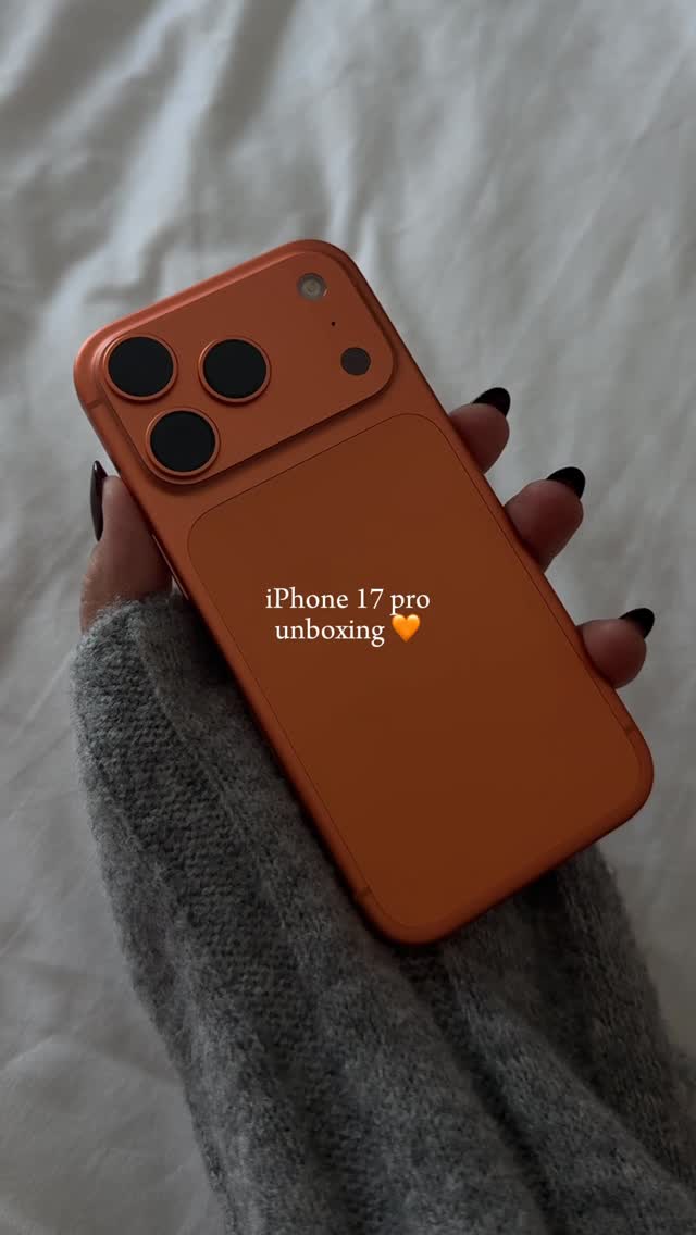 A hand holding an orange iPhone, the cover image is soft and blurry, with the phone’s camera array and the person’s black nail manicured hand being in focus. The phone is held against a white, textured background. The phone is a bright orange color with black cameras, and the fingers have very well-maintained and shiny black nails.