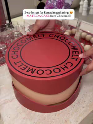 A person's hands are holding a large, round red box. The box has the words "Chocomelt" in a circular pattern around the top. Inside the box, the curved edge of a Matilda cake is visible with its light color. A white tag with text is at the top. The cake is on a light-colored surface.