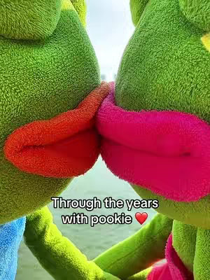 The image features two green plush toys with large, contrasting mouths. One toy's mouth is vibrant red, while the other is hot pink. They are positioned close together as if kissing or nuzzling, with their mouths nearly touching. The background isn't clear, but the plush toys are the center focus. The green bodies provide a soft texture and the image is aimed to be cute.