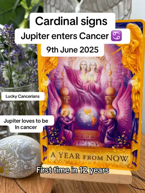 The image is a tarot card with text overlayed on top. The card is predominantly yellow and gold with illustrations of figures with wings. The text is in a white box with black text. There is a small vase of lavendar in the left corner. There is a polished stone resting on what look to be a piece of wood.