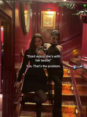 Two young women are standing on a staircase, posing for a photo. The background has red walls and a warm lighting. The woman in the foreground is wearing a black dress and holding a phone, while the other is wearing a black halter dress. Overlayed text appears on the image, and the composition suggests they are at a party or a social gathering.