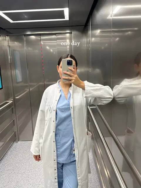A person in a white lab coat and light blue scrubs takes a mirror selfie inside what appears to be an elevator. The interior is metallic silver, with a prominent "one day" text overlay.
